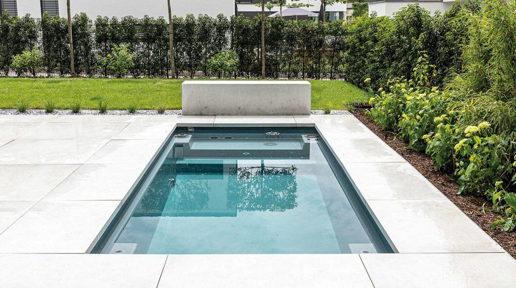 Moderner rechteckiger Pool mit klar blauem Wasser, umgeben von großen weißen Fliesen und grünem Garten mit Büschen und Bäumen