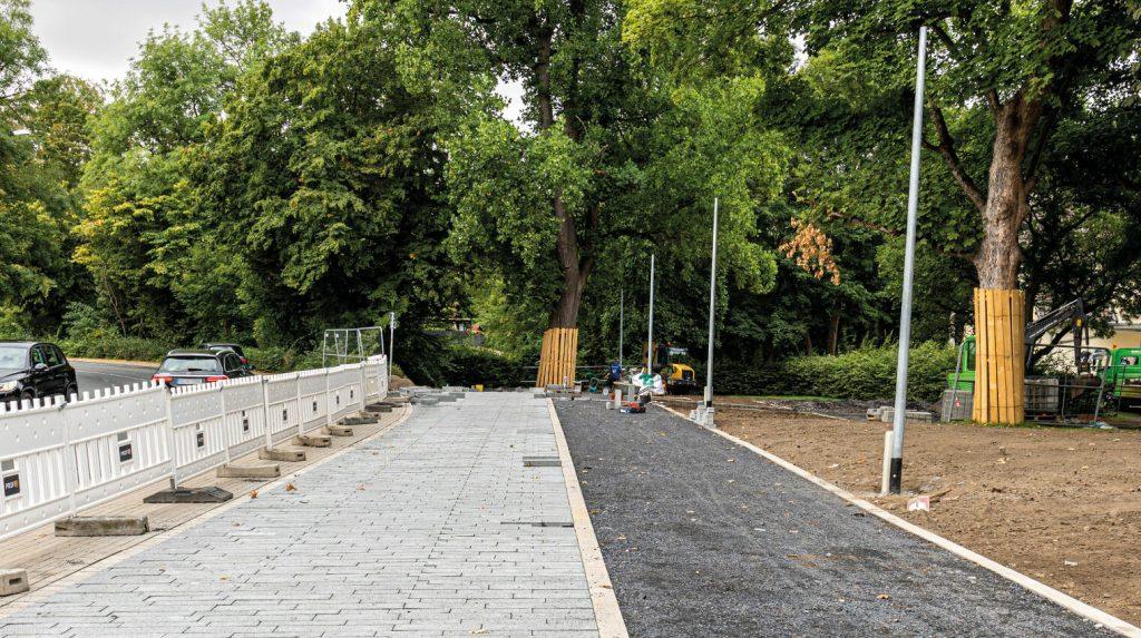 Baustelle eines Gehwegs mit neu verlegten grauen Pflastersteinen und daneben unbefestigtem Weg, umgeben von Bäumen und Baustellenabsperrungen