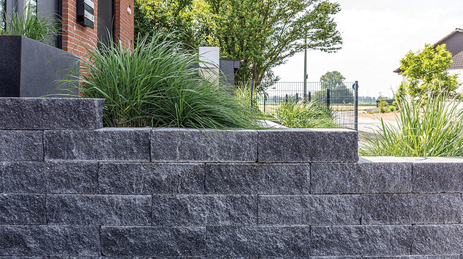 Graue Steinmauer als Grundstücksbegrenzung mit grünen Pflanzen dahinter und einem Metallzaun im Hintergrund