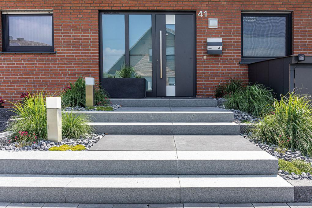 Moderner Hauseingang mit grauen Stufen, zwei quadratischen Laternen, Pflanzbeeten mit Ziergräsern und rotem Backsteinfassade