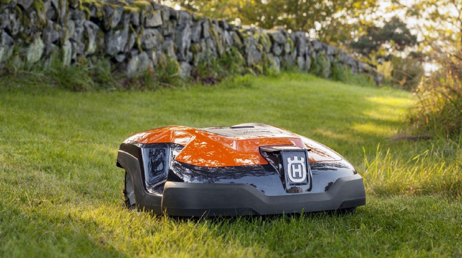 Oranger Mähroboter mit schwarzem Unterteil auf grünem Rasen vor einer Steinmauer im Garten