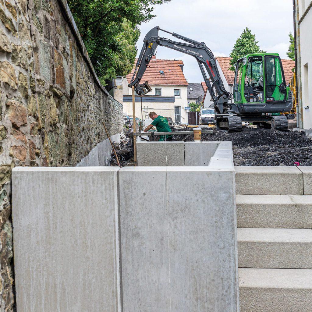 Bauarbeiter mit Bagger arbeiten an einem Garten- und Landschaftsbauprojekt neben einem Gebäude mit Treppen aus Betonplatten