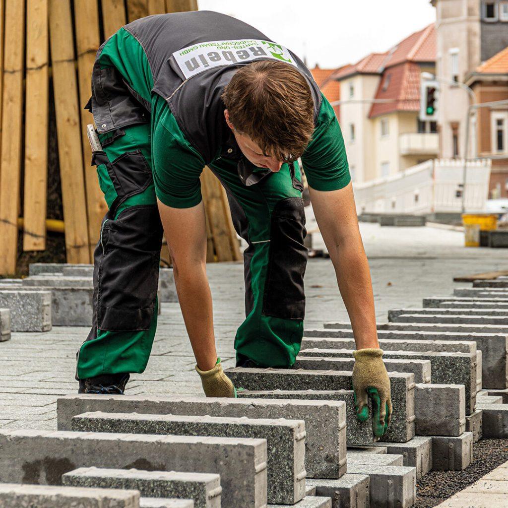 Mann in Arbeitskleidung verlegt graue Pflastersteine auf einem Gehweg in einer Stadt