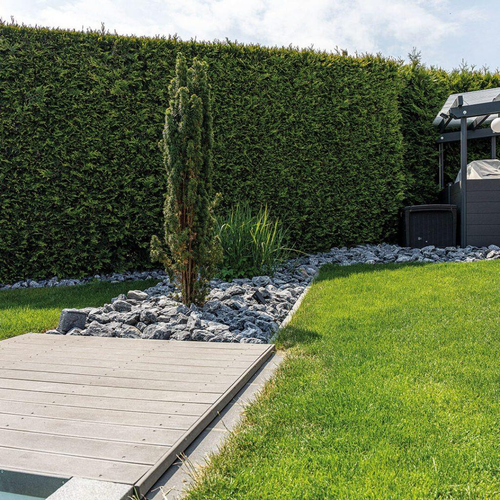 Gepflegter Garten mit grünem Rasen, Steinen und einer Hecke im Hintergrund, daneben eine Holzterrasse