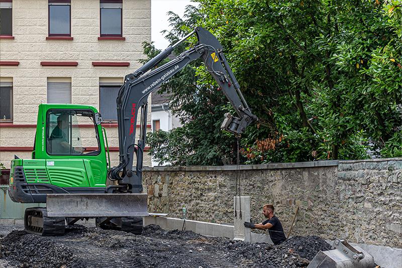 Grüner Bagger hebt Betonteile an einer Baustelle neben einer Steinmauer, Arbeiter führt Arbeiten im Erdreich aus