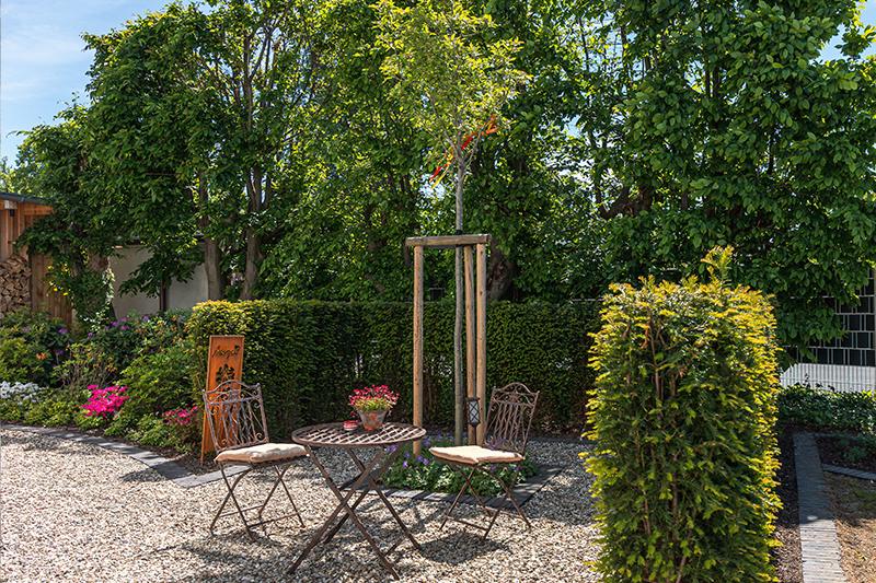 Kleiner Sitzbereich mit zwei Stühlen und rundem Tisch auf Kies, umgeben von grünen Büschen und Bäumen, mit einem Blumentopf auf dem Tisch