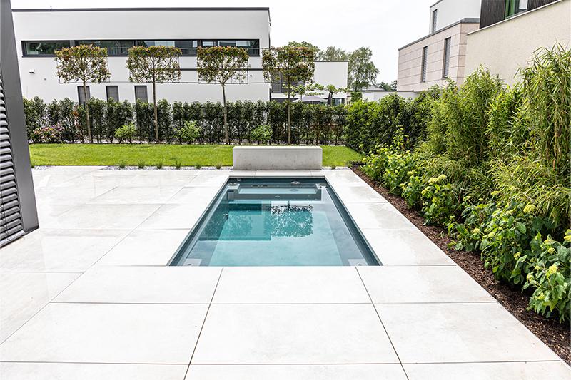 Moderner rechteckiger Pool mit klar blauem Wasser, umgeben von großen weißen Fliesen und grünem Garten mit Büschen und Bäumen