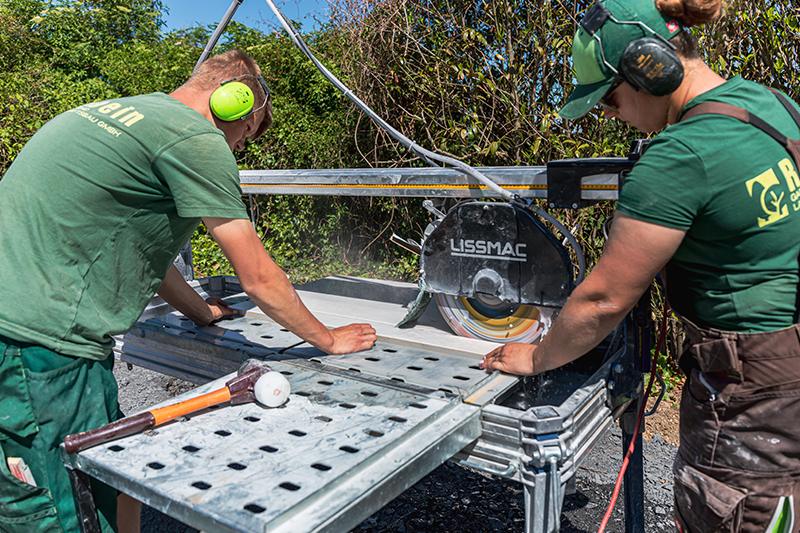 Zwei Männer schneiden mit einer Lissmac-Steintrennmaschine große Platten auf einer Baustelle, beide tragen Gehörschutz und grüne Arbeitskleidung