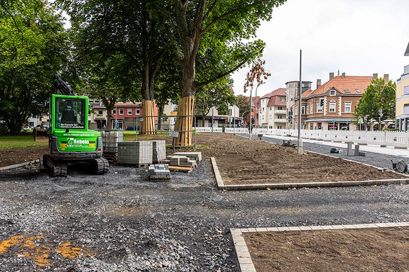 Baustelle eines Gehwegs mit neu verlegten grauen Pflastersteinen und daneben unbefestigtem Weg, umgeben von Bäumen und Baustellenabsperrungen