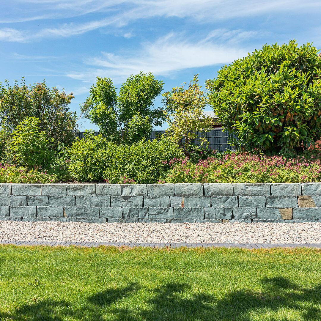 Steinmauer mit grünen Sträuchern und Blumen dahinter, Kiesweg davor, grüner Rasen im Vordergrund unter blauem Himmel