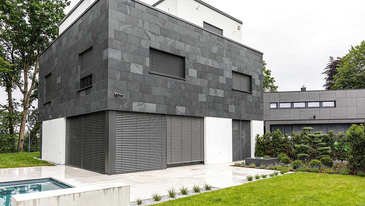 Moderne Bauhaus-Villa mit grauer Schieferfassade im Obergeschoss und weißen Wänden im Erdgeschoss, umgeben von Garten und Terrasse