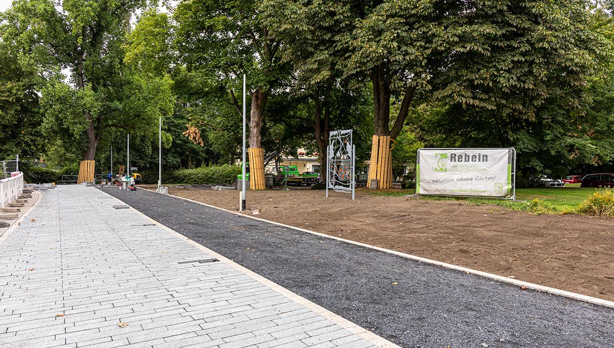 Baustelle eines Gehwegs mit neu verlegten grauen Pflastersteinen und daneben unbefestigtem Weg, umgeben von Bäumen und Baustellenabsperrungen