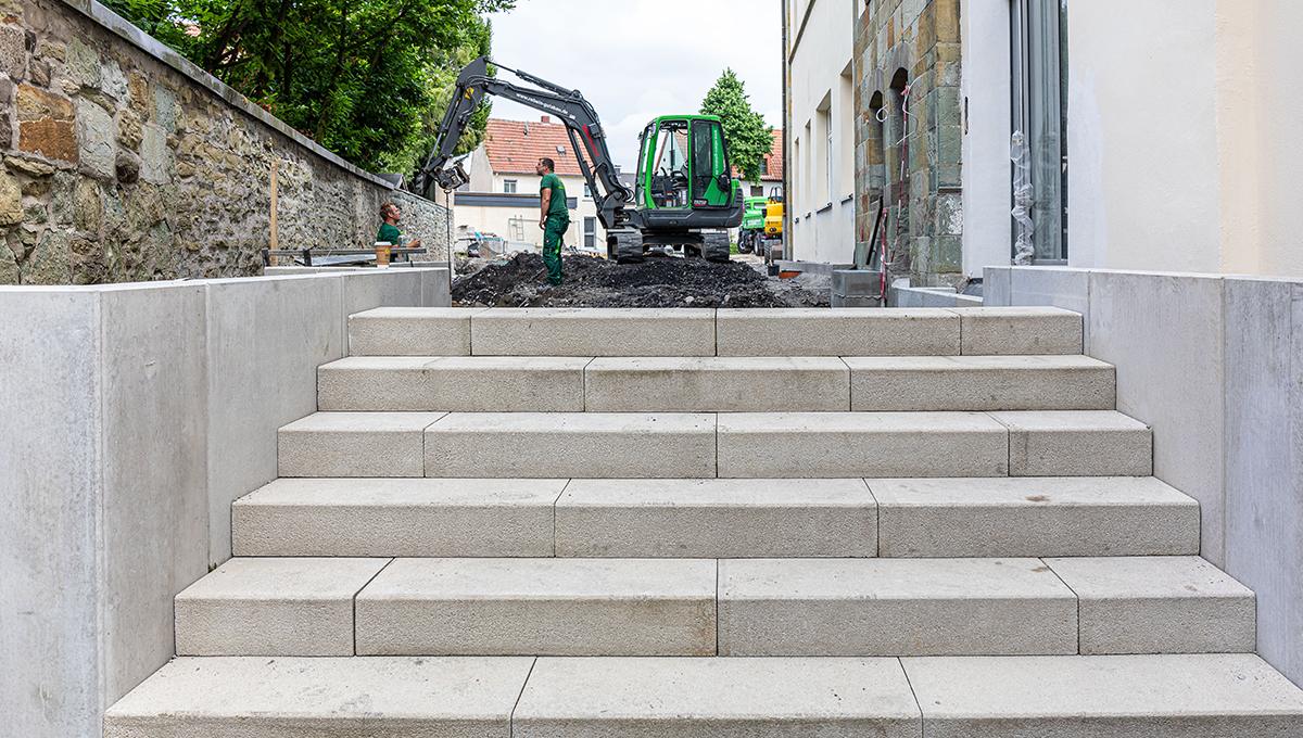 Breite Treppe aus hellen Betonstufen zwischen zwei Mauern, im Hintergrund zwei Bauarbeiter und ein Bagger vor einem Gebäude