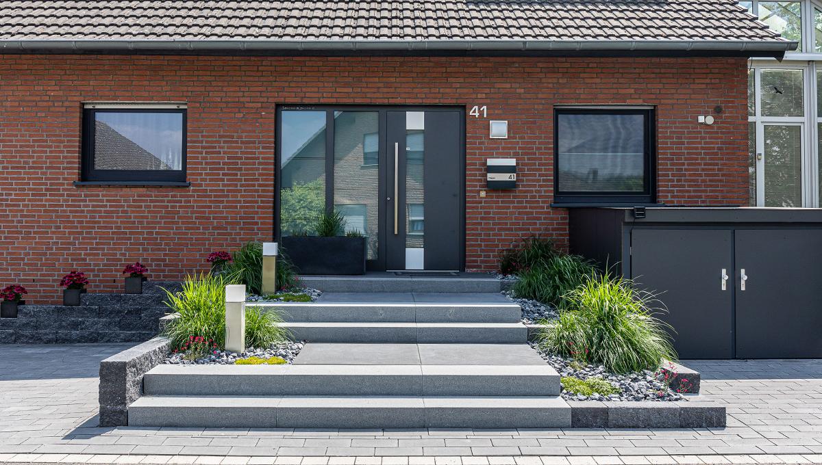 Moderner Hauseingang mit grauen Stufen, zwei quadratischen Laternen, Pflanzbeeten mit Ziergräsern und rotem Backsteinfassade