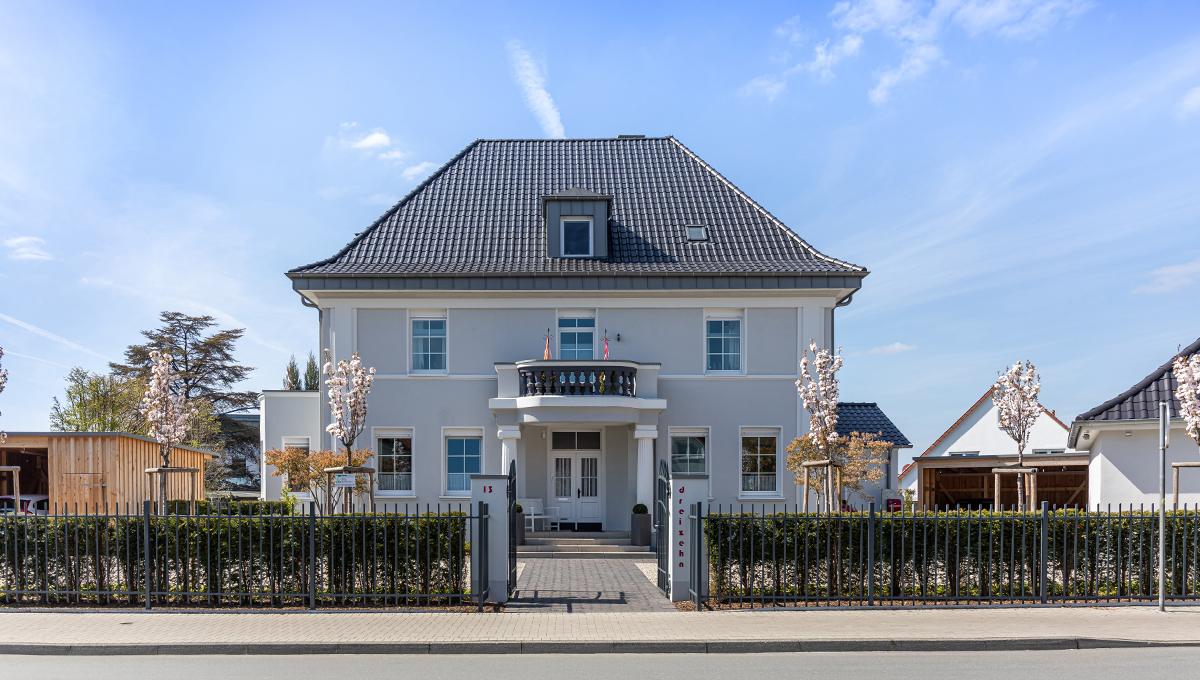 Großes zweistöckiges Wohnhaus mit grauem Dach und weißer Fassade, umgeben von einem schwarzen Metallzaun und kleinen blühenden Bäumen