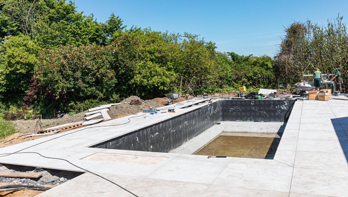 Baustelle eines rechteckigen Pools mit teilweise gefülltem Wasser und umliegender Terrasse aus großen hellen Platten, zwei Arbeiter im Hintergrund