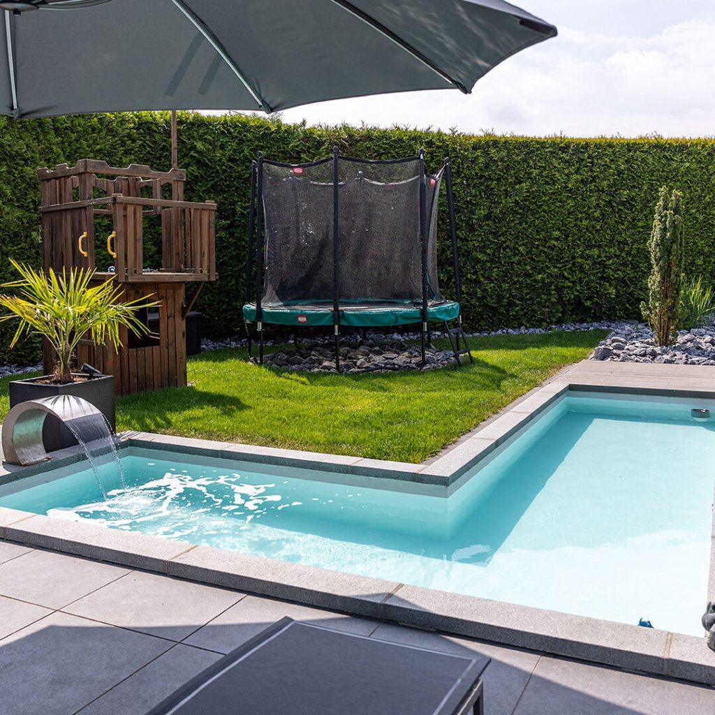 Moderner Eckpool mit Wasserfall, umgeben von Rasen, Kinderspielhaus und Trampolin im Garten