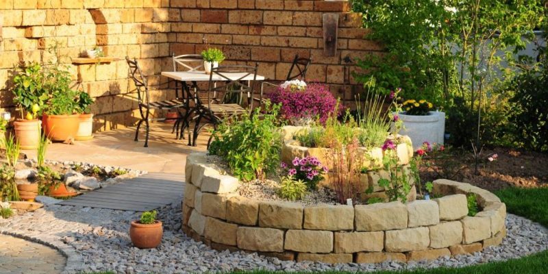 Kräuterspirale aus Sandstein mit verschiedenen Pflanzen und Blumen, umgeben von Kies und Rasen, neben einer Terrasse mit Tisch und Stühlen