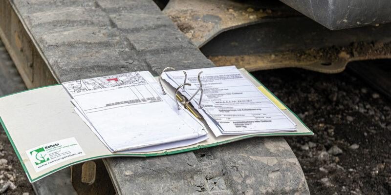 Offener Ordner mit Dokumenten liegt auf der Kette eines Baumaschinenfahrwerks auf Erde