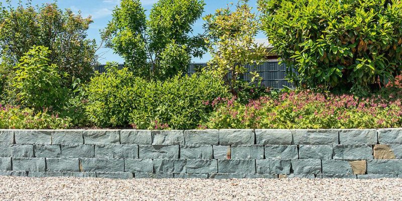 Rebein_Referenzen_Galeriebilder_villa_mauer_kiesweg_etagen-1080×1080 Steinmauer mit grünen Sträuchern und Blumen dahinter, Kiesweg davor, grüner Rasen im Vordergrund unter blauem Himmel