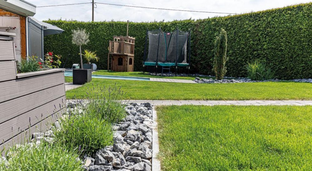 Rebein_gartengestaltung_Blogbeitrag_Rasenpflege Gepflegter Garten mit grünem Rasen, Lavendelpflanzen, Holzspielhaus, Trampolin und Hecke im Hintergrund