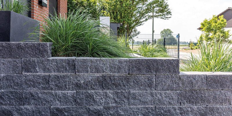 Graue Steinmauer als Grundstücksbegrenzung mit grünen Pflanzen dahinter und einem Metallzaun im Hintergrund